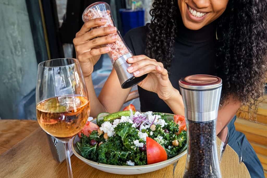A woman eating a salad is holding a salt grinder with a glass of wine and a pepper grinder on the table